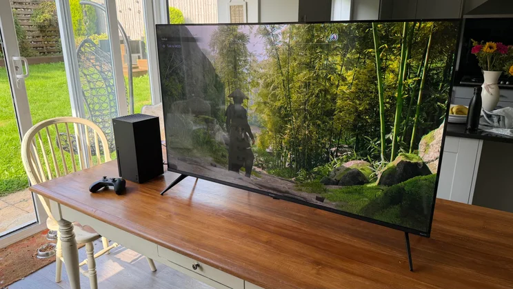 Wide front view of the Sharp HJ6725KB Roku TV set up on a wooden dining table next to an Xbox Series X console, displaying video game graphics