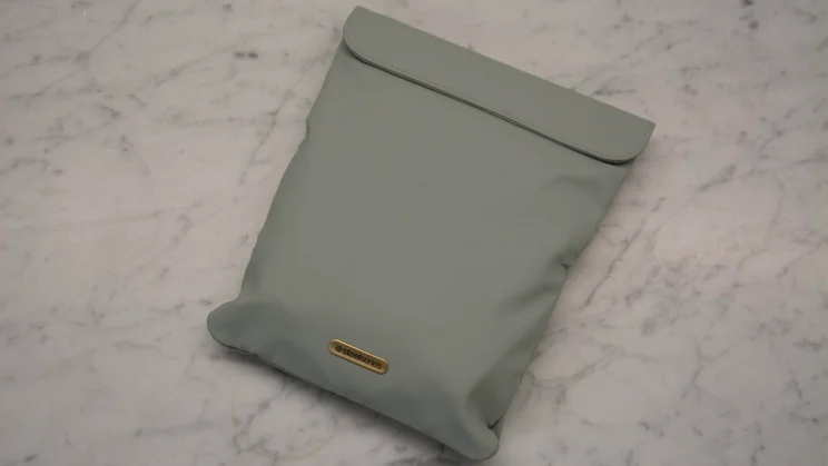 A sage green protective pouch for the SteelSeries Arctis Nova Elite gaming headset, featuring a top fold-over flap and a gold metal logo badge near the bottom on a marble surface