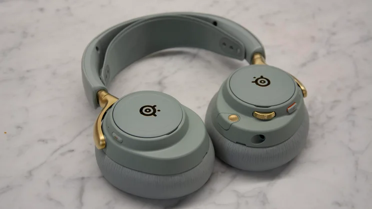 The SteelSeries Arctis Nova Elite gaming headset in sage green with matte gold accents on the hinges, featuring earcups with gold logos resting on a marble surface