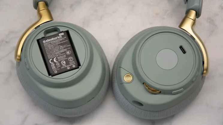 A close-up of the SteelSeries Arctis Nova Elite gaming headset with the outer plate removed from one earcup to reveal the removable battery compartment alongside the opposite earcup's controls