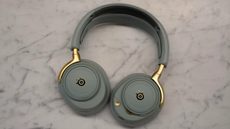The sage green and gold SteelSeries Arctis Nova Elite gaming headset lying flat on a marble surface, displaying the gold logo backplates and earcup controls