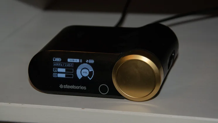 Front view of the black and gold GameHub wireless transmitter unit for the SteelSeries Arctis Nova Elite gaming headset, displaying an active OLED screen with volume level indicators and connection status