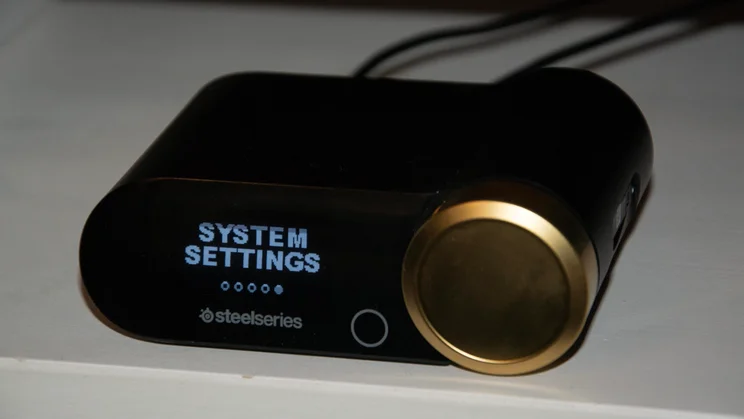 Close-up of the GameHub base station for the SteelSeries Arctis Nova Elite gaming headset with the screen illuminated to read "SYSTEM SETTINGS" alongside the gold control dial