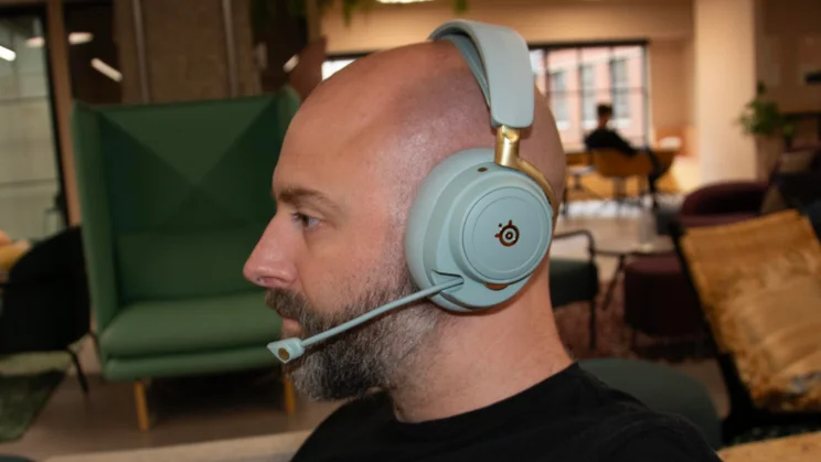 A side profile of a man wearing the sage green SteelSeries Arctis Nova Elite gaming headset with its flexible boom microphone extended toward his mouth