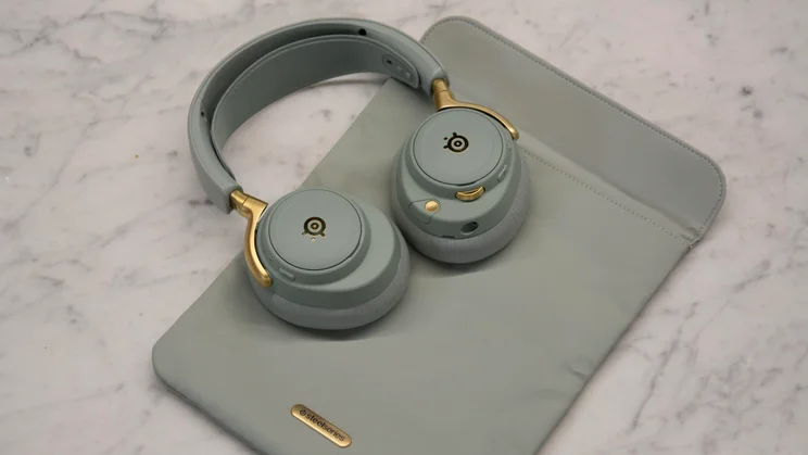 The SteelSeries Arctis Nova Elite gaming headset in sage green with gold accents resting on top of its matching fabric carrying pouch on a marble tabletop