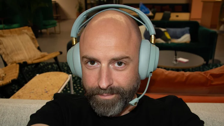A front-facing selfie of a man wearing the SteelSeries Arctis Nova Elite gaming headset in sage green, showing the fit of the headband and retractable boom microphone