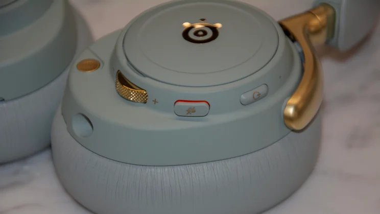 Close-up view of the left earcup controls on the SteelSeries Arctis Nova Elite gaming headset, highlighting the gold volume dial, microphone mute button, power button, and 3.5mm audio port