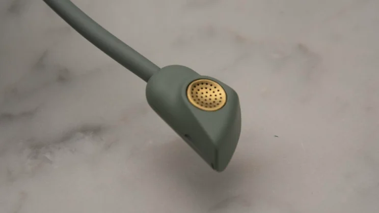 Detailed macro shot of the tip of the retractable boom microphone on the SteelSeries Arctis Nova Elite gaming headset, showing the sage green housing and perforated gold grille