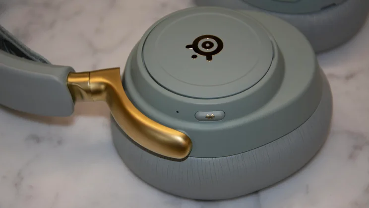 A close-up view of the right earcup on the SteelSeries Arctis Nova Elite gaming headset, showing the Bluetooth pairing button alongside the matte gold headband arm and backplate logo