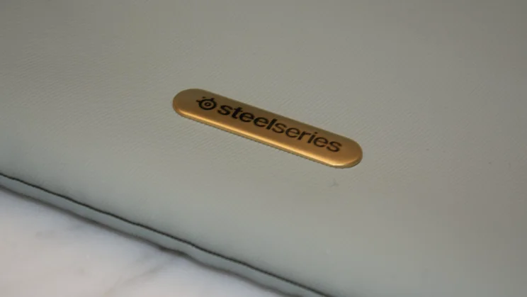 A close-up shot of the raised metallic gold SteelSeries logo badge affixed to the sage green fabric of the SteelSeries Arctis Nova Elite gaming headset's carrying pouch