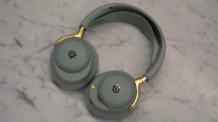 A top-down view of the sage green SteelSeries Arctis Nova Elite gaming headset lying flat on a light grey marble surface, showcasing its matte gold hinges and backplate logos