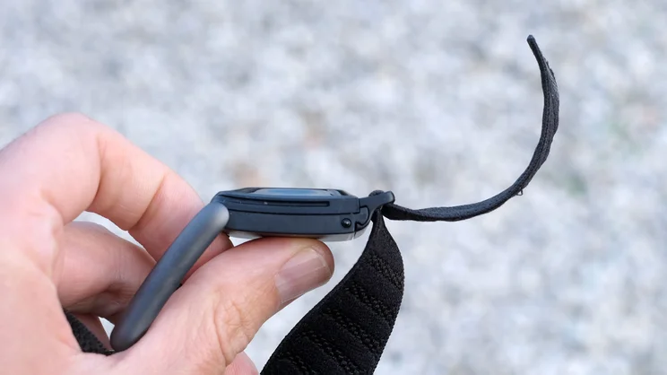 Whoop 5.0 held in the hand with the band unfastened, showing the pod's side profile and the loose end of the band