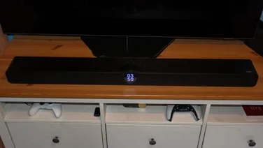Front view of the black WiiM Bar soundbar resting on a wooden TV console unit below a television, showing its central circular digital clock display