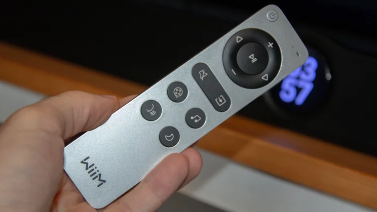 A hand holding the silver and black wireless remote control for the WiiM Bar soundbar