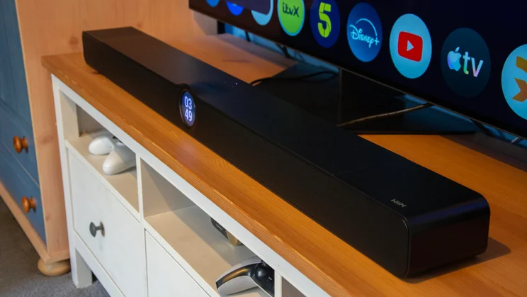 Side-angled view of the black WiiM Bar soundbar resting on a wooden TV console in front of a smart TV screen showing streaming apps