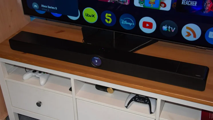 Front view of the WiiM Bar soundbar positioned directly in front of a TV, with its circular display illuminated with a volume level icon